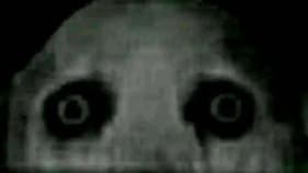 A face, particularly a set of eyes looking up at the viewer, appearing to be partially concealed. It's eyes are just dark sockets from which the eye ball should be with a faint white ring in the center of the socket to imitate an iris 